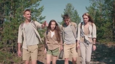Sırt çantalı dört genç kadın ve erkeğin güneşli yaz gününde ormanda birlikte yürüyüş ve gülümsemelerini izliyoruz.