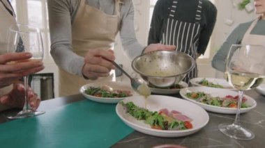 Aşçılık kursunda diğer insanlar birlikte yemek masasının etrafında durup izlerken, yemek pişirme kursunda hazırlanmış restorana benzeyen sosları üzerine döken, tanınmayan bir adamın yavaş bir orta bölüm çekimi.