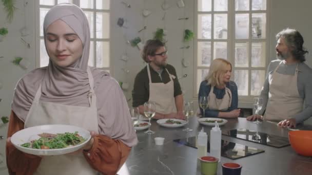 身着头巾和围裙的年轻而美丽的穆斯林女子慢镜头的Pan中期肖像 拿着美味的菜品站在烹饪课上 坐在厨房桌上的人在后台交谈 — 图库视频影像