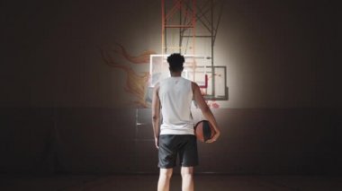 Dolly-in portresi. Dikiz manzaralı erkek basketbolcu. Kapalı alandaki sahada spot ışığı altında duruyor.