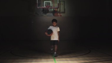 Elinde topla kameraya doğru yürüyen ve spot ışıklarıyla karanlık basketbol sahasında poz veren küçük bir çocuğun yavaş yavaş portresi.