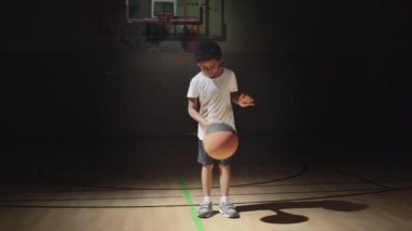 Yavaş çekimde, karanlık bir basketbol sahasında spot ışıkları altında duran yakışıklı bir çocuk ve top sürüyor.