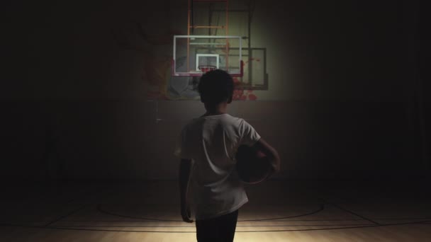 聚光灯照射在小男孩身上的慢镜头后跟随着球在黑暗的室内篮球场上向篮筐走去 — 图库视频影像