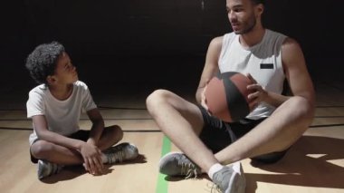 Spor giyim mağazasında elinde top tutan ve koyu renk basketbol sahasında oturan neşeli siyahi adamın el kamerasıyla çekilmiş fotoğrafı. Gülümsüyor ve oğluyla konuşuyor.