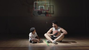 Genç siyah basketbolcunun el kamerasıyla çekilmiş bir fotoğrafı. Kapalı sahada yerde oturuyor ve çocuğa sayı atmayı öğretiyor.