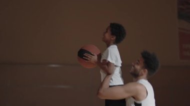 Genç Afro-Amerikan oyuncunun yavaş çekimde gülümsemesi ve oğlunu kaldırarak kapalı sahada basketbol oynamasına yardım etmesi.