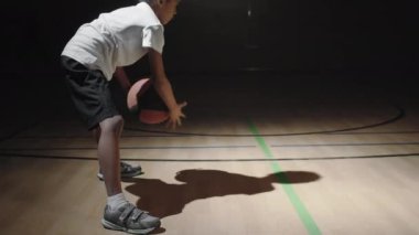Siyah çocuğun spor giyim kuşağındaki yavaş el kamerasıyla karanlık basketbol sahasında top sürerken, spot ışıkları üzerinde parlarken.
