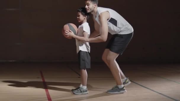 スローモトラッキングショットの幸せな黒選手でスポーツウェア持ち上げる彼の小さな息子と彼を助けるシュートバスケットボールにフープで屋内裁判所 — ストック動画