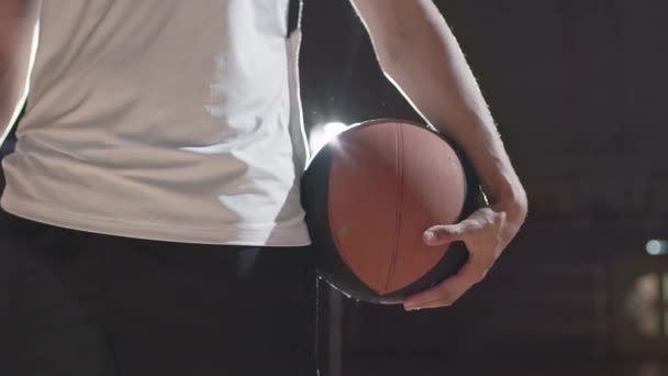 闭合的中段镜头 缓慢跟踪无法辨认的篮球运动员 球站在黑暗的室内场地上 背景明媚 — 图库视频影像