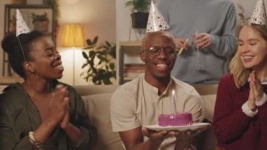 Parti şapkası takmış, erkek arkadaşları doğum günü pastasında mum üflerken alkışlayan ve tezahürat yapan genç arkadaşların PAN çekimi.