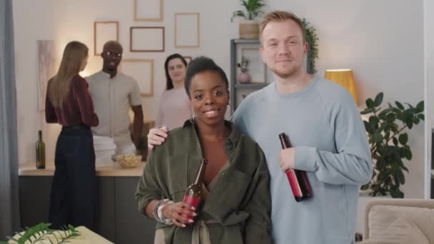 アパートの自宅パーティーでカメラに笑顔でビールボトルを持った若い黒人女性と陽気な男のポートレートPanのショット 3人の友人が背景でチャット — ストック動画