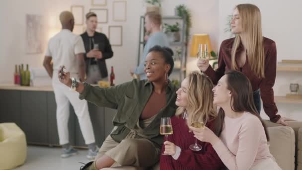 在公寓的沙发上坐着一杯香槟或白葡萄酒 并在家庭聚会上自拍的快乐年轻女性的中景照片 三个男人在后面聊天 — 图库视频影像