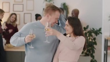 Beyaz şarapla dolu kadehler tutan ve ev partisinde dans eden mutlu genç kadın ve adamın orta boy fotoğrafı.