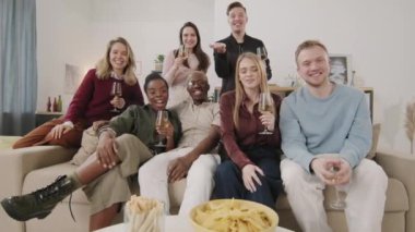 Beyaz şarapla dolu kadehlerle dolu bir grup mutlu arkadaş kanepede oturmuş video konuşmalarında sohbet ediyorlar. El sallayıp gülüyorlar.