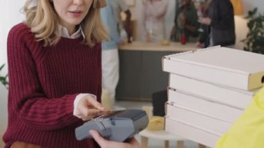 Teslimatçının elinde tuttuğu banka kartına dokunan ve pizza parasını dairesinde ödeyen mutlu genç bir kadın. Arka planda sohbet eden arkadaş grubu