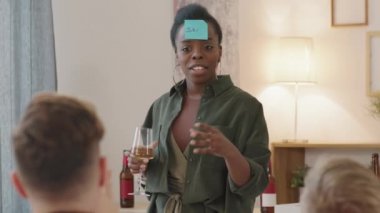 Alnında yapışkan bir notla kayağın elinde beyaz şarap kadehi tutarak arkadaşlarıyla birlikte tahmin oyunu oynarken sorular sorduğu orta boy mutlu siyah bir kadın fotoğrafı.