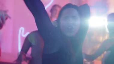 Mor ışıklandırmalı bir odada arkadaşlarıyla parti yaparken kameranın önünde dans eden hevesli genç bir kadının orta boy fotoğrafı.