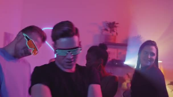 ネオンのサングラスを身につけた青年とその仲間たちが カメラの前で紫色の照明で踊っている 若い女性Djバックグラウンドで — ストック動画