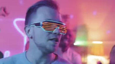 Ev partisinde DJ 'le kamera önünde güneş gözlüklü genç adamın neon çubuğu ya da ışık dansı ve şarkı söylerken yakın çekim görüntüsü.