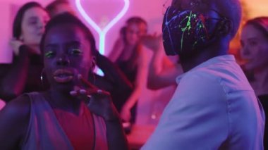 Kirpiklerinde yeşil neon maskaralı genç siyahi kadın ve partide mor ışıkla dans eden yüz maskeli Afro-Amerikalı adam. Arka planda kadın DJ 'liği