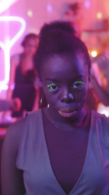 Canlı makyajlı, halka küpeli ve kameraya gülümseyen genç siyahi bir kadının dikey görüntüsü.