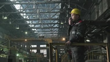 Sarı başlıklı beyaz erkek bitki denetçisinin telsizle konuşurken ve aktif olarak hareket eden çalışanları kontrol ederken orta görüş görüntüsü.