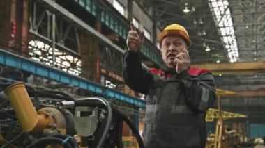 Sarı başlıklı Asyalı erkek fabrika yöneticisinin orta boy görüntüsü. Telsizle konuşuyor ve çalışanlara hareket eden makineleri kontrol ediyor.