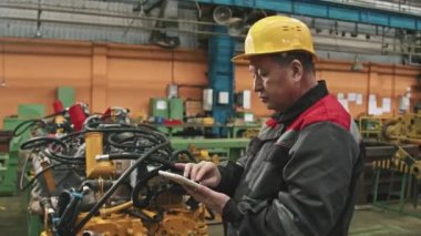 Büyük traktör fabrikasının montaj atölyesinde dijital tablet kullanan sarı şapkalı erkek bitki yöneticisinin orta boy görüntüsü.