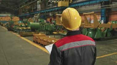 Büyük traktör fabrikasının montaj atölyesinde yürürken sarı başlıklı erkek bitki denetleyicisinin dikiz görüntüsü.