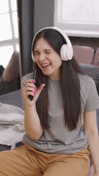 垂直ショットの精力的な若い女性でヘッドフォンでソファに家に座って音楽にジャミング — ストック動画