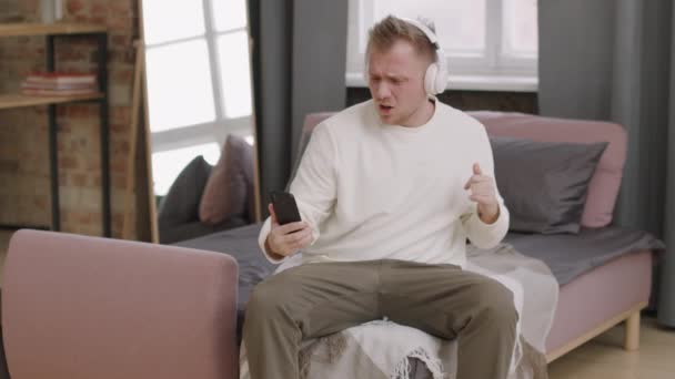 Średni Ujęcie Szczęśliwego Młodego Człowieka Słuchawkach Siedzi Kanapie Patrzy Telefon — Wideo stockowe