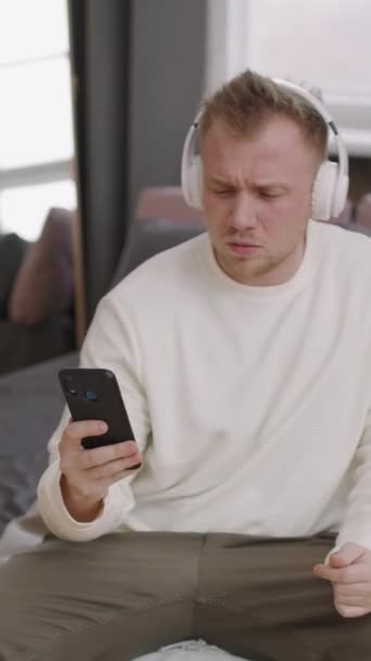 Вертикальный Снимок Счастливого Молодого Человека Наушниках Сидящего Диване Слушающего Музыку — стоковое видео