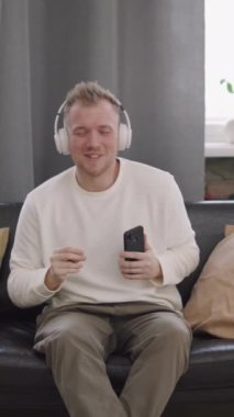 Dikey çekimde, kulaklıklı yakışıklı genç adam elinde cep telefonuyla kanepede otururken müzik eşliğinde dans ediyor.