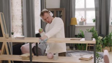 Evde rafları temizlerken müzik eşliğinde şarkı söyleyen ve dans eden kulaklıklı yakışıklı bir gencin orta boy fotoğrafı.