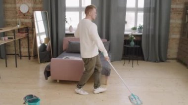 Genç adamın dans ederken ve müziğin keyfini çıkarırken dairesinin yerlerini paspaslarken orta boy fotoğrafı.