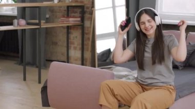 Kulaklıklı mutlu genç bir kadının cep telefonuyla müzik dinlerken kanepede oturup gülümsemesi.