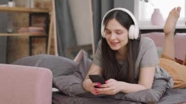 Kulaklıklı genç bir kadının cep telefonuyla sosyal medyada gezinirken kanepede dinlenip müzik dinlerken PAN görüntüsü.