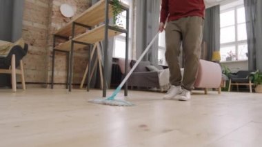 Düşük açılı, tanınmayan bir gencin dairede yerleri paspaslarken ve müzikle dans ederken görüntüsü.