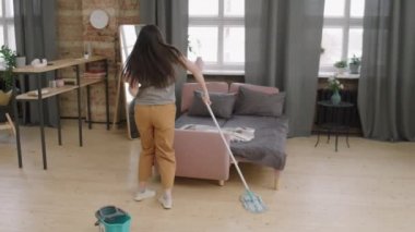 Dairesinde müzik dinlerken dans eden ve yerleri paspaslayan mutlu genç bir kadının orta boy fotoğrafı.