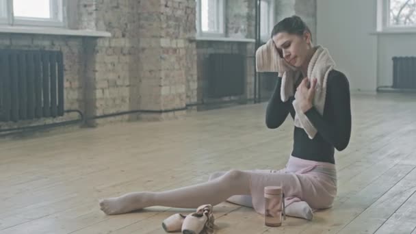 年轻优雅的芭蕾舞演员坐在木制地板上 在空荡荡的舞蹈班里 经过了艰苦的芭蕾舞训练 凝视着窗户后 休息了下来 — 图库视频影像