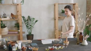 Orta boy PAN portresi. Yavaşça gülümseyen kadının el yapımı doğal kozmetikler için çeşitli malzemelerle masada duran kameraya bakması.