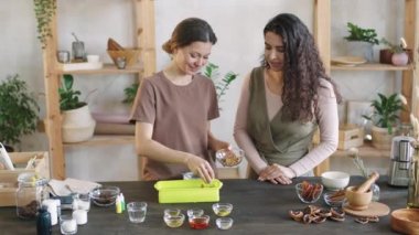 İki gülümseyen kadının el yapımı doğal organik sabunu kurutulmuş çiçek yapraklarıyla süslediği ve kişiselleştirdiği orta büyüklükte bir PAN fotoğrafı.