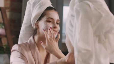 Çekici genç bir kadın ve en yakın arkadaşının yüz maskesi takarak birbirlerine güzellik rutin prosedürleri uygulayarak omuz üstünden orta bir çekim yapıyorlar.