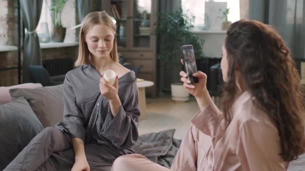 20多岁的年轻漂亮姑娘们坐在舒适的床上 用智能手机录下一段视频 记录她们日常的美容美发 并在她们的皮肤上涂上乳霜和润肤液 — 图库视频影像
