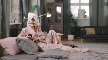 PAN 'da, pembe ipek pijamalı, akıllı telefon kullanan ve altın nemlendirici çarşaf maskeli genç bir kadın resmi.