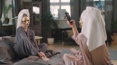 Yüzü altın sarısı maskeli güzel bir kadının akıllı telefon kamerasına poz verip kadın arkadaşıyla yatakta ipek pijama giyip eğlendiği orta boy bir fotoğraf.