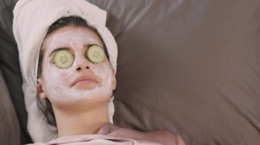 Yüzünde kil maske ve gözlerinde salatalık dilimleri ile yatakta yatarken gülümseyen beyaz bir kadının en iyi görüntüsü.