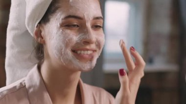 Genç, gülümseyen bir kadın ve en yakın arkadaşının birlikte güzellik prosedürleri uygulayarak yüzlerine kilden maske takmalarına yakından bakın.