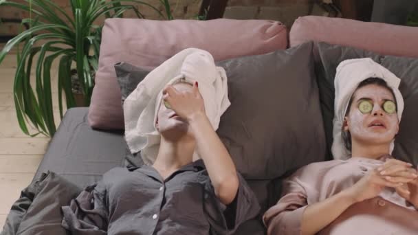 上ビューPanショットの2人の若い女性の親友横たわっベッドの上でマスクを顔に適用され キュウリスライス上の目チャットとゴシップ — ストック動画