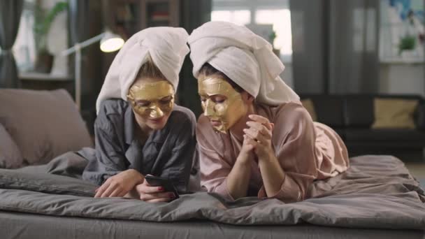 Pan中镜头 慢慢地 年轻漂亮的女人躺在舒适的床上 头戴时髦的金床单面具 看着智能手机 — 图库视频影像